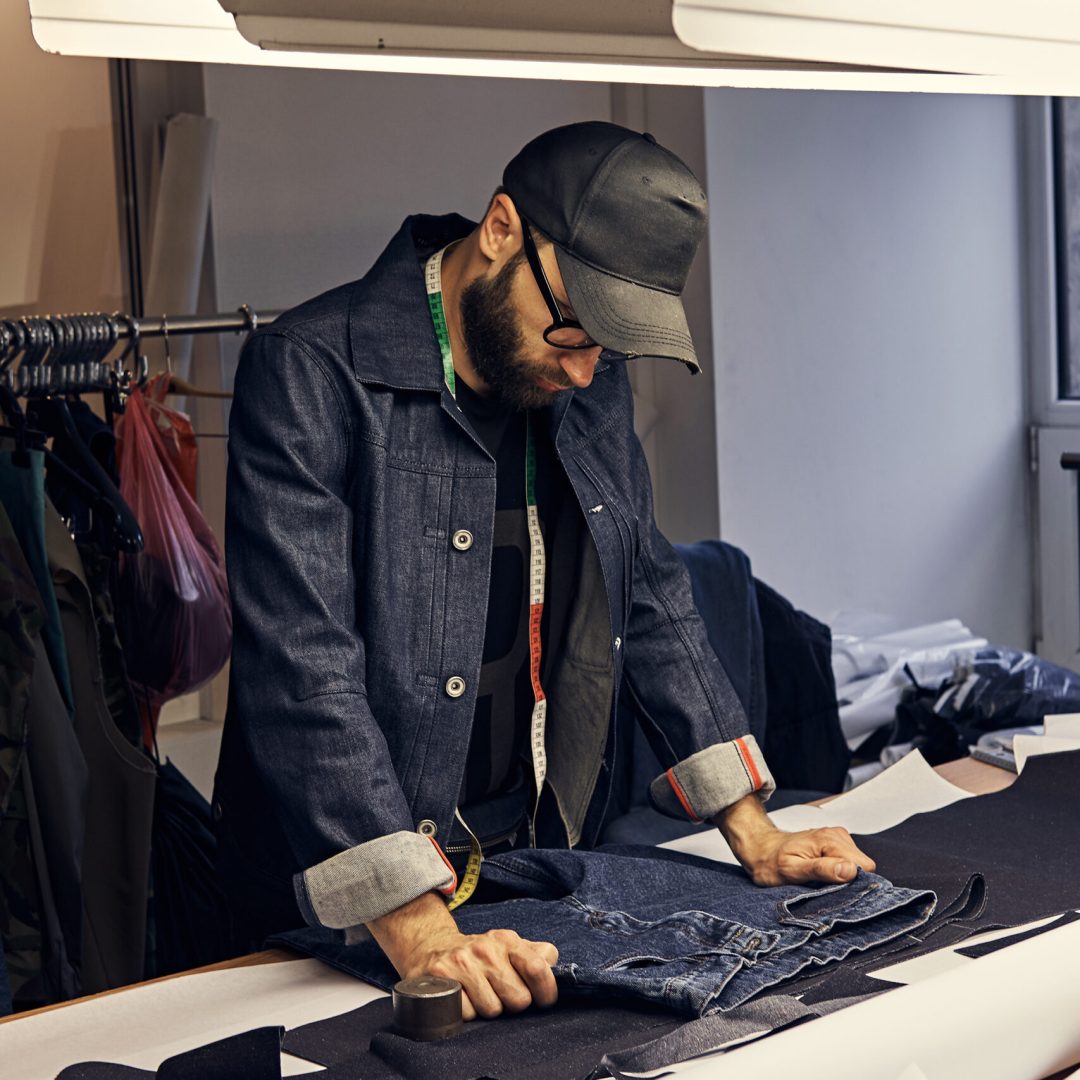 Handsome bearded tailor working with jeans material at a sewing workshop.